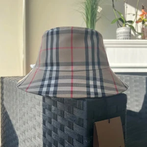 Rutig bucket hat från Burberry - Snygg bucket hat från Burberry i klassiskt beige, svart, vitt och rött rutmönster. Hatten har bred brätte och är tillverkad i bomull. Perfekt accessoar för att lyfta din outfit med en ikonisk streetstyle-vibe.