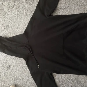 Svart hoodie med huva och ficka - En klassisk svart hoodie med huva och snörning. Tröjan har en stor magficka framtill och långa ärmar.