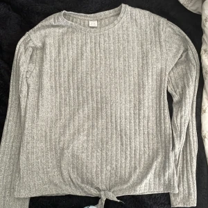 Grå ribbad tröja med knyt från Lindex - Grå långärmad tröja från Lindex i storlek 170. Tröjan är ribbad och har en snygg knytning längst ner framtill. Perfekt för dig som gillar en enkel men trendig stil. Mjuk och skön att ha på sig.