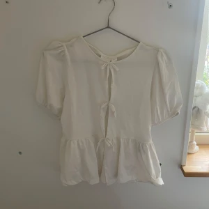 Vit blus med knyt från Gina Tricot - Supersöt vit blus från Gina Tricot med puffiga korta ärmar och tre knyt framtill. Blusen har volang nedtill och är gjord i ett lätt och luftigt material, perfekt för en fräsch och trendig look. Passar dig som gillar romantiska detaljer och vill ha något som sticker ut.strl 158/164. Använd en gång.