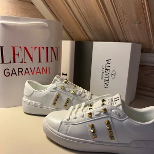 Valentino Rockstud vita sneakers - Snygga vita Valentino Rockstud sneakers i skinn med ikoniska guldiga nitar längs sidorna och på tungan. Klassisk rund tå, platt sula och coola detaljer som ger en lyxig vibe. Perfekta för dig som vill sticka ut med stilrena men edgy skor.