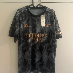 Arsenal bortatröja Adidas svart/guld - Snygg Arsenal bortatröja från Adidas med geometriskt mönster i svart och grått. Guldiga detaljer som klubbmärke, Adidas-logga och sponsortryck. Tillverkad i lätt och ventilerande material, perfekt för match eller träning.