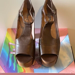 Bruna platåklackar från Jeffrey Campbell - Snygga bruna klackskor i skinn från Jeffrey Campbell med kraftig platåsula och hög klack i trälook. Skorna har öppen tå, rundad form och T-rem med spänne. Perfekta för dig som vill sticka ut med en retrovibe och cool siluett.