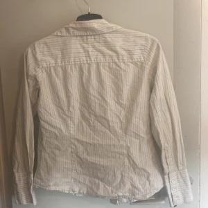 Vit randig skjorta från Clara - Vit skjorta från Clara med tunna ljusgula och grå ränder. Klassisk krage, långärmad och knäppning framtill. Tillverkad i mjuk bomull som känns skön mot huden. Perfekt för dig som gillar en clean och stilren look.