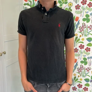 Svart pikétröja från Ralph Lauren - Klassisk svart pikétröja från Ralph Lauren med röd broderad logga på bröstet. Tröjan har korta ärmar, krage med knappar och är tillverkad i mjuk bomull. Perfekt till jeans eller shorts för en clean och stilren look.