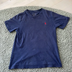 Marinblå t-shirt från Polo Ralph Lauren - Klassisk marinblå t-shirt från Polo Ralph Lauren med röd broderad logga på bröstet. T-shirten har rund halsringning och korta ärmar. Tillverkad i mjuk bomull som känns skön mot huden. Perfekt för en clean och stilren look.