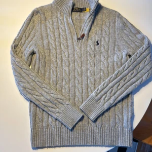 Ralph lauren half zip kabelstickad - Kabelstickad grå tröja från Polo Ralph Lauren med halv dragkedja och klassisk broderad logga på bröstet. Tröjan har hög krage, ribbade muddar och är långärmad. Perfekt för alla årstider och du kan matcha den till vilken outfit som helst, hör av dig vid minsta intresse!