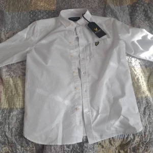 Vit skjorta från Lyle & Scott, stl S - Klassisk vit skjorta från Lyle & Scott i storlek S. Skjortan har button down-krage, lång ärm och snygg broderad logga på bröstet. Tillverkad i bomull och har knappar framtill. Perfekt för en clean och stilren look.