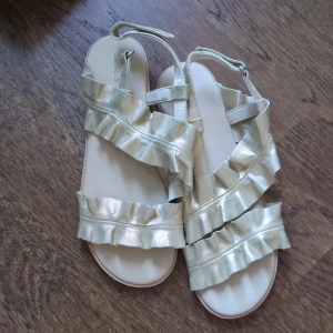 Guldiga sandaler från Zara - Helt nya sandaler från Zara. Platt sula och öppen tå gör dem perfekta för varma dagar. Justerbar rem runt hälen för bra passform. Aldrig använda.
