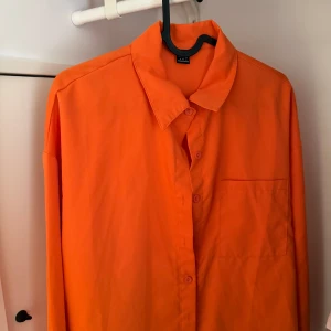Orange långärmad skjorta med ficka - Snygg orange skjorta med klassisk krage och knäppning framtill. Skjortan har en bröstficka och långa ärmar, perfekt för dig som vill sticka ut med färg. Enkel och stilren design som passar till många olika outfits. Använd endast en gång 