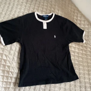 Svart t-shirt Polo Ralph Lauren - Svart t-shirt från Polo Ralph Lauren med vit kontrastkrage och vita detaljer vid ärmslut. Klassisk logga broderad på bröstet. Skön passform och tillverkad i mjuk bomull. Perfekt för en clean och stilren look. Passar XS/S dam