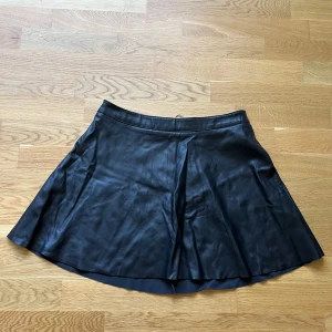 Svart skinnkjol med dragkedja - Snygg svart kortkjol i skinnimitation med en silverfärgad dragkedja framtill. 