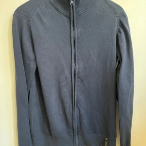 Mörkblå kofta från Indicode - Mörkblå kofta från Indicode med hel dragkedja framtill och ribbad krage. Tröjan är långärmad och har en stilren, enkel design utan mönster. 