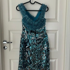 Glittrig blå långklänning från Invito - Snygg långklänning från Invito i glittrande blått och silver med paljetter över hela klänningen. Överdelen har volangiga axelband och omlott-detalj. Tillverkad i polyester, nylon och lycra. Perfekt för dig som vill sticka ut med en unik look!