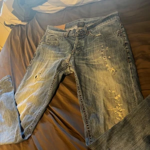 Blå slitna jeans från Dondup - Säljer ett par blå jeans från Dondup med slitningar och hål både fram och bak. Jeansen har klassisk femficksdesign, normal passform och är tillverkade i bomull. Perfekt för dig som gillar en rå och avslappnad streetstil. W31 L28/29