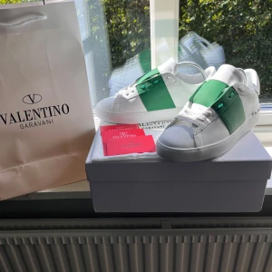 Valentino Garavani sneakers  - Snygga Valentino Garavani sneakers i vitt skinn med en bred grön rem över mitten och coola nitar på hälen. Klassisk rund tå, vita snören och exklusiv känsla. Perfekta för dig som vill sticka ut med stilrena detaljer och lyxig design. Dem är helt nya och allting som finns med på bild ett följer med.                      Hör av er om ni har några frågor!😊