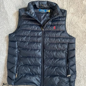 Svart dunväst från Polo Ralph Lauren - Snygg svart dunväst från Polo Ralph Lauren med quiltad design och röd broderad logga på bröstet. Västen har dragkedja framtill och två praktiska fickor med dragkedja. Perfekt att slänga över en hoodie för en chill look.