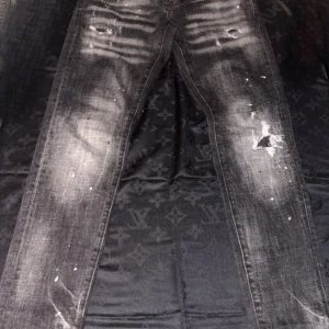 Svarta Dsquared2 jeans med slitningar - Säljer ett par svarta jeans från Dsquared2 med coola slitningar och blekta detaljer. Jeansen har en smal passform och är dekorerade med vita färgstänk för en edgy look. Klassisk femficksmodell med Dsquared2-logga vid fickan.