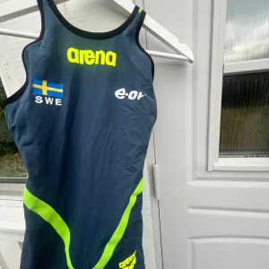 Arena tävlingsbaddräkt  SWE - tävlingsbaddräkt från Arena (landslagskollektion) med neongula detaljer och svensk flagga. Använd på tävlingar, bra skick. Endast eon markeringen som har åkt av lite