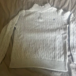Ljusgrå stickad tröja från Polo Ralph Lauren - Kabelstickad ljusgrå tröja från Polo Ralph Lauren med halv dragkedja och broderad marinblå logga på bröstet. Tröjan har ribbade muddar, hög krage och lång ärm. Perfekt för lager-på-lager och snygg till jeans. Storlek 18-20 på ralph lauren som motsvara ungefär 170-188cm beror på hur tajt eller baggy man vill ha det.