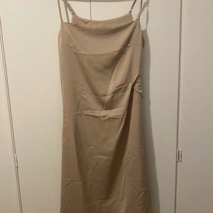 Beige medellångklänning med smala axelband - Snygg beige medellångklänning med smala axelband och enkel, stilren design. Klänningen har en lätt glansig yta och rak passform som faller snyggt längs kroppen. Perfekt för dig som gillar minimalistisk stil och vill ha en klänning som känns både trendig och tidlös. Klänningen är lite skrynklig men måste vara strykas.