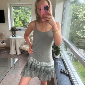 Grågrön kort klänning med spets - Supersöt grågrön kort klänning med tunna axelband och vacker spetsdetalj nertill. Klänningen har en avslappnad passform och spetsen ger en romantisk touch. Perfekt för varma dagar när du vill känna dig både chill och snygg.