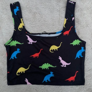 Svart crop top med färgglada dinosaurier - En svart croppad linnetopp från SHEIN med färgglada dinosaurie-tryck i blått, grönt, gult, rött, rosa och lila. Toppen har breda axelband och ärmlös design, perfekt för dig som gillar lekfulla och unika plagg. Materialet är mjukt och stretchigt för en bekväm passform.