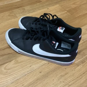 Svarta Nike sneakers med vit swoosh - Snygga svarta Nike sneakers med klassisk vit swoosh på sidan och vit sula med svart rand. Skorna har snörning och rund tå, tillverkade i syntetmaterial och textil. Perfekta för dig som gillar en clean och sportig look.