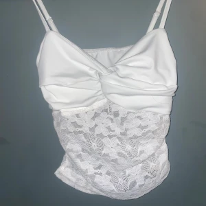 Vit spetsig linnetopp  - Supersöt vit linnetopp med smala axelband och en stor rosett framtill. Nedre delen är i blommig spets som ger en romantisk vibe. Toppen är figurnära och perfekt för dig som vill sticka ut med en feminin och trendig look.