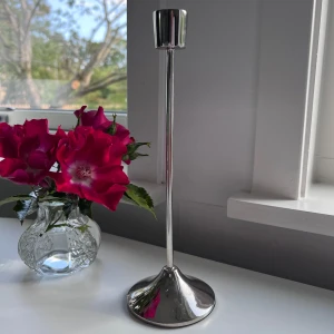 Silvrig ljusstake - Silverfärgad ljusstake i metall från Rusta. Den har en modern, stilren design med rund fot och hög smal hals. Perfekt snygg detalj i inredningen. Säljer pga att den inte kommer till användning. Den är 25cm hög och foten är 8cm bred. 🤍