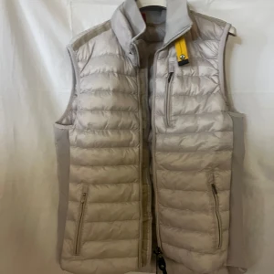 Beige dunväst från Parajumpers - Snygg beige dunväst från Parajumpers med quiltad design och hög krage. Västen har dragkedja framtill, tre fickor med dragkedja och den klassiska gula Parajumpers-detaljen vid halsen. Perfekt lager-på-lager-plagg för kyliga dagar.