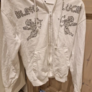 Vit hoodie från Blinglucid med strass - Vit hoodie från Blinglucid med dragkedja, huva och stora fickor fram. Framsidan har coola strassdetaljer med texten 'BLING LUCID' och två änglar i glittriga stenar. Materialet är mjuk bomull och polyester, perfekt för dig som gillar statement-plagg.