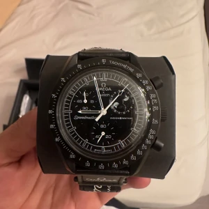 Säljer en helt ny och oanvänd SwatchMission to the MoonPhase Bioceramic MoonSwatch.  - - Endast öppnad för foto. - Plasten är kvar.  - Tillverkningsår: 2024  Skriv gärna om du har ytterligare frågor avseende klockan.   Information gällande klockan: Klockan är en del av Swatchs begränsade upplaga av MoonSwatch-kollektionen, inspirerad av Omega Speedmaster. Den har en svart keramisk boett och armband, en svart urtavla med en månfasindikator och en tachymeter-skal. Klockan drivs av en kvartsrörelse och är vattentät till 30 meter.