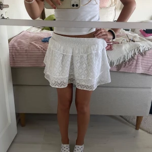 Vit volangkjol från Gina Tricot - Supersöt vit kortkjol från Gina Tricot med bred smock i midjan och volangdetalj. Kjolen har ett diskret geometriskt mönster i tyget och är gjord i ett lätt, luftigt material. Perfekt för varma dagar och enkel att styla med crop top eller linne. Kjolen är i storlek 158/164 och passar xxs-s jättebra!💗