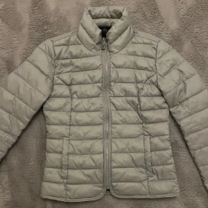 Ljusgrå pufferjacka från Only, S - Säljer en ljusgrå pufferjacka från Only i storlek S. Jackan har quiltad design, hög krage och dragkedja framtill. Perfekt för kyliga dagar och enkel att matcha med olika outfits. Snygg och stilren look som funkar året runt.