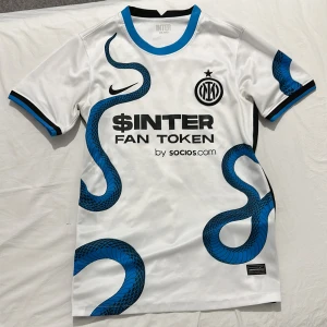 Inter Milan vit bortatröja Nike - Inter Milans vita bortatröja från Nike med blå och svart ormtryck som slingrar sig över framsidan. Tröjan har klubbmärke, sponsorloggor och blå detaljer vid krage och ärmslut. Tillverkad i lätt och ventilerande material, perfekt för match eller träning.