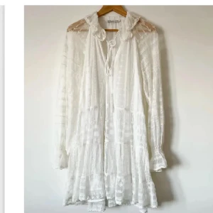 Vit spetsklänning med volang - Superfin vit kortklänning från Zara med romantiska spetsdetaljer, volangkant och knytning vid halsen. Långärmad och luftig med transparenta partier och v-ringning. Perfekt för sommarens alla tillfällen.