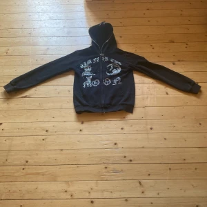 Svart hoodie Aim For The Moon XS - Svart hoodie med huva från Aim For The Moon i storlek XS. Framsidan har glittriga stenar och tryck med texten 'AIM FOR THE MOON' samt coola motiv. Dragkedjan har en unik metallberlock. Perfekt för dig som gillar streetstyle och detaljer.