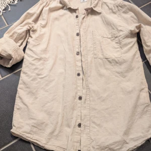 Beige skjorta i bomull/linne från C, stl 152 - Snygg beige skjorta från C i storlek 152. Skjortan är tillverkad i mjuk bomull och linne, har klassisk krage, bröstficka och knäppning framtill. Perfekt för en avslappnad och stilren look. Långärmad modell som är lätt att matcha med det mesta.