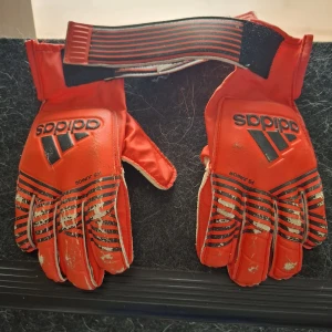 Röda Adidas målvaktshandskar strl 6 - Säljer ett par röda Adidas målvaktshandskar i juniorstorlek med svarta ränder och logga. Handskarna har bred kardborreknäppning och insida i ljust syntetmaterial för bra grepp. Perfekta för fotbollsträning eller match!