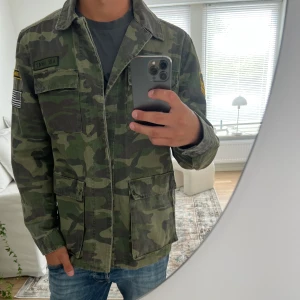 Camouflage overshirt med patchar från Adrian hammond  - Säljer en grön camouflage overshirt med coola patchar och bröstfickor. Jackan har klassisk krage, knäppning framtill och stora fickor nertill. Perfekt för dig som vill ha en streetig och avslappnad look.