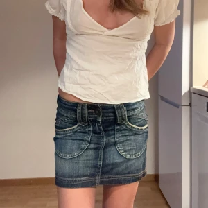 Lågmidjad kort jeanskjol - Snygg kort jeanskjol i blå denim med markerade sömmar och stora, rundade fickor framtill. Midjemått: 37cm, Längd: 32,5cm, Storlek: 32 (xs/s)