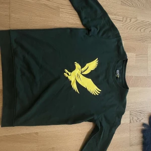 Grön Lyle & Scott tröja med gul fågel - Mörkgrön långärmad tröja från Lyle & Scott med stor gul fågel-logga på bröstet. Klassisk rund halsringning och skön passform. Perfekt för dig som gillar stilrena plagg med en tydlig logga. Materialet är mjuk bomull.