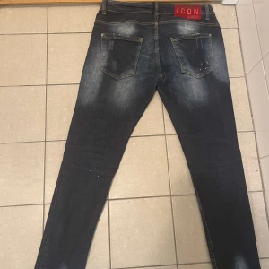 Mörkblå distressed jeans ICON - Säljer ett par mörkblå jeans från ICON med slitna detaljer, patchar och färgstänk framtill. Jeansen har fem fickor, normal passform och klassisk knappgylf. Snyggt tvättade partier och röd logotyp bak i midjan ger extra edge.