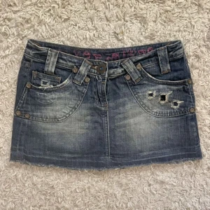 Blå jeanskjol med slitningar - Snygg blå jeanskjol med slitna detaljer och rå kant nertill. Klassisk femficksmodell med bälteshällor och coola nitar. Bak finns en läderpatch med ros-motiv. Perfekt för dig som gillar en edgy och avslappnad stil. Köp via Swish + frakt (ej köp nu) eller direkt via vinted @moa.06 (pga plicks nya avgifterk) 🩷