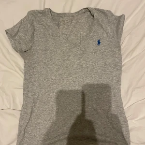 Grå v-ringad t-shirt från Polo Ralph Lauren - Snygg grå t-shirt från Polo Ralph Lauren med v-ringning och korta ärmar. Klassisk blå logga broderad på bröstet. Skön och mjuk bomullskvalitet, perfekt till jeans eller shorts. Enkel och stilren design som alltid funkar.