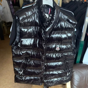 Svart dunväst från Moncler - Snygg svart dunväst från Moncler med glansig finish och klassisk logga på bröstet. Västen har hög krage, två dragkedjefickor och stängs med både dragkedja och tryckknappar. Perfekt för lager-på-lager-stil under kyliga dagar.