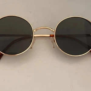 Runda solglasögon med guldfärgade bågar - Snygga runda solglasögon med tunna guldfärgade metallbågar och mörka linser. Skalmarna har bruna detaljer längst ut för en extra touch. Perfekt accessoar för dig som vill sticka ut med en retrovibe.