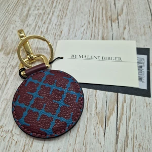 By Malene Birger KeyKey nyckelring - Rund nyckelring från By Malene Birger i vinrött "skinn" med blått mönster på ena sidan och präglad logga på andra. Guldiga metalldetaljer och karbinhake gör den enkel att fästa på väska eller nycklar. Perfekt accessoar för att lyfta din look.