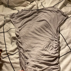 Ljusgrå ribbad topp med rynkade sidor - Superskön ljusgrå topp med korta ärmar och snygga rynkade detaljer längs sidorna. Modellen är figurnära och passar perfekt till jeans eller kjol. Tunt och mjukt bomullsmaterial som känns bekvämt hela dagen.